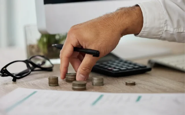 Persona apilando monedas en un escritorio junto a una calculadora, gafas y documentos