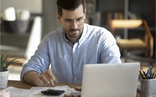Hombre con gafas usando una calculadora y un portátil en un escritorio con papeles